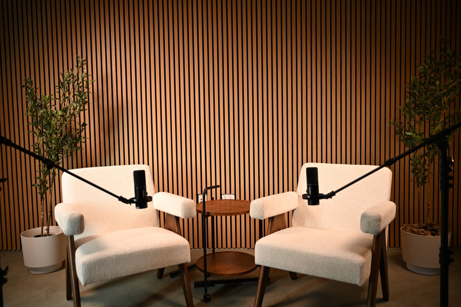 Wide angle of the Axeom podcast studio in Addison, TX, showing two boucle armchairs, mic booms, and a wood-slat backdrop.